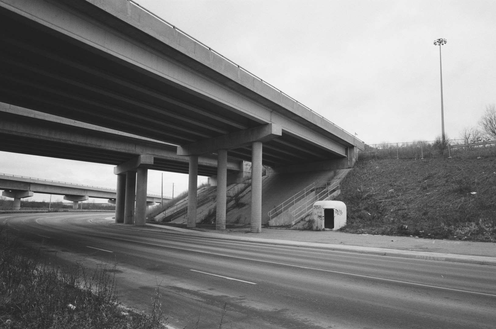 A black & white photograph of a mysterious hut underneith a highway underpass