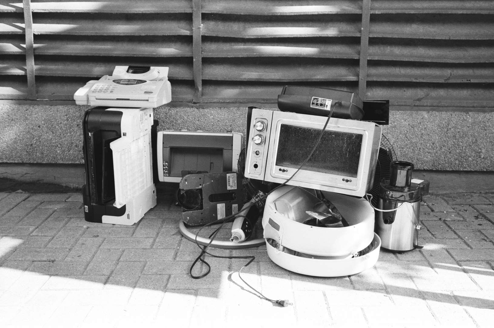 A black and white photograph of home appliances left on the sidewalk: a fax machine, printer, toaster oven, a juicer, and a fan.