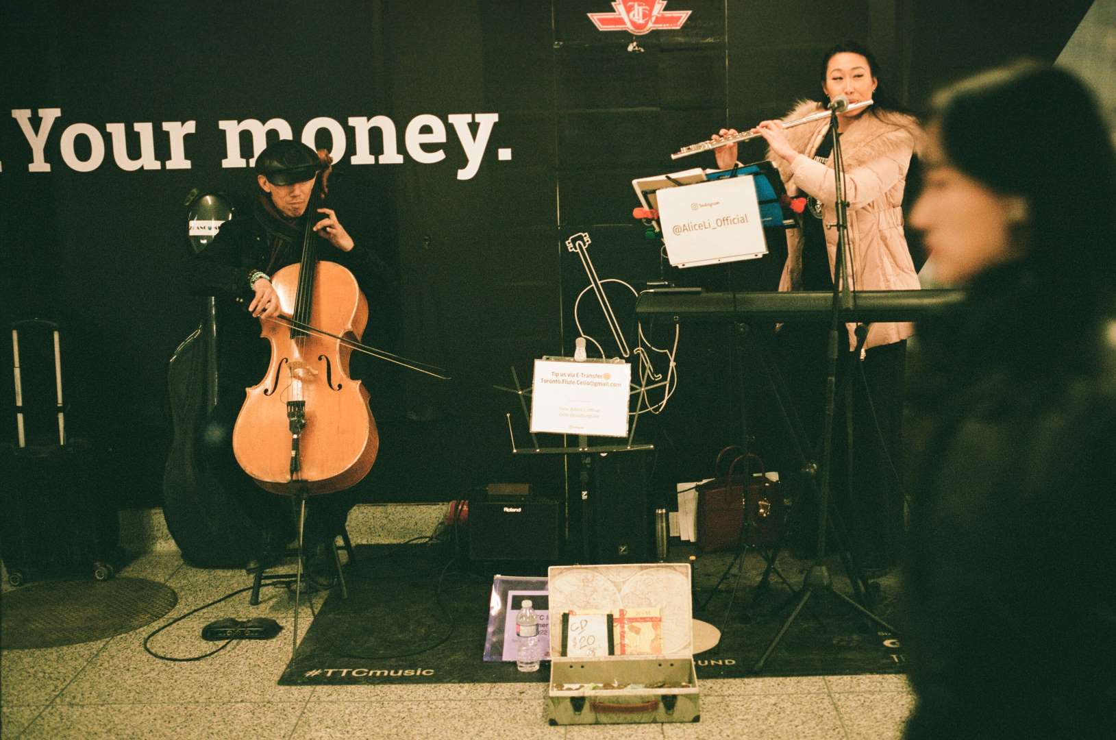 A colour photograph of two buskers, the woman playing flute, the man playing cello, in the Bloor-Yonge Subway.