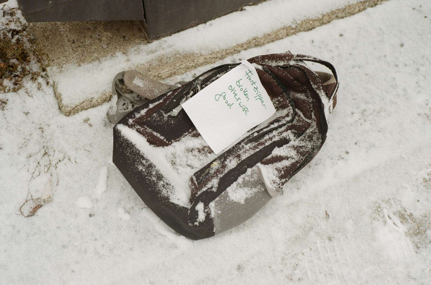 A colour photograph of a backpack left in the snow with a note 'front zipper broken, otherwise good - free'