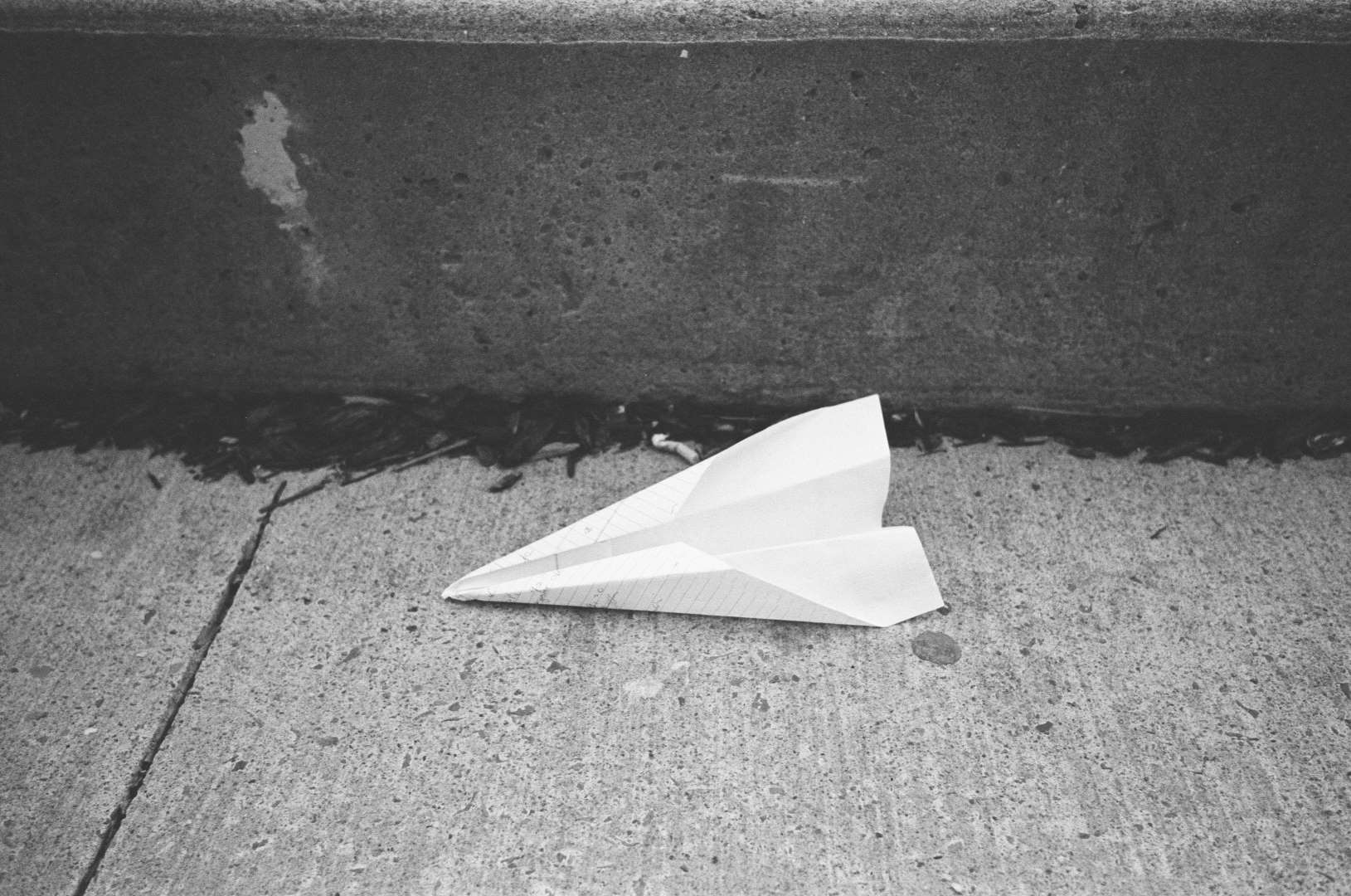 A black and white photograph of a paper airplane left on the sidewalk