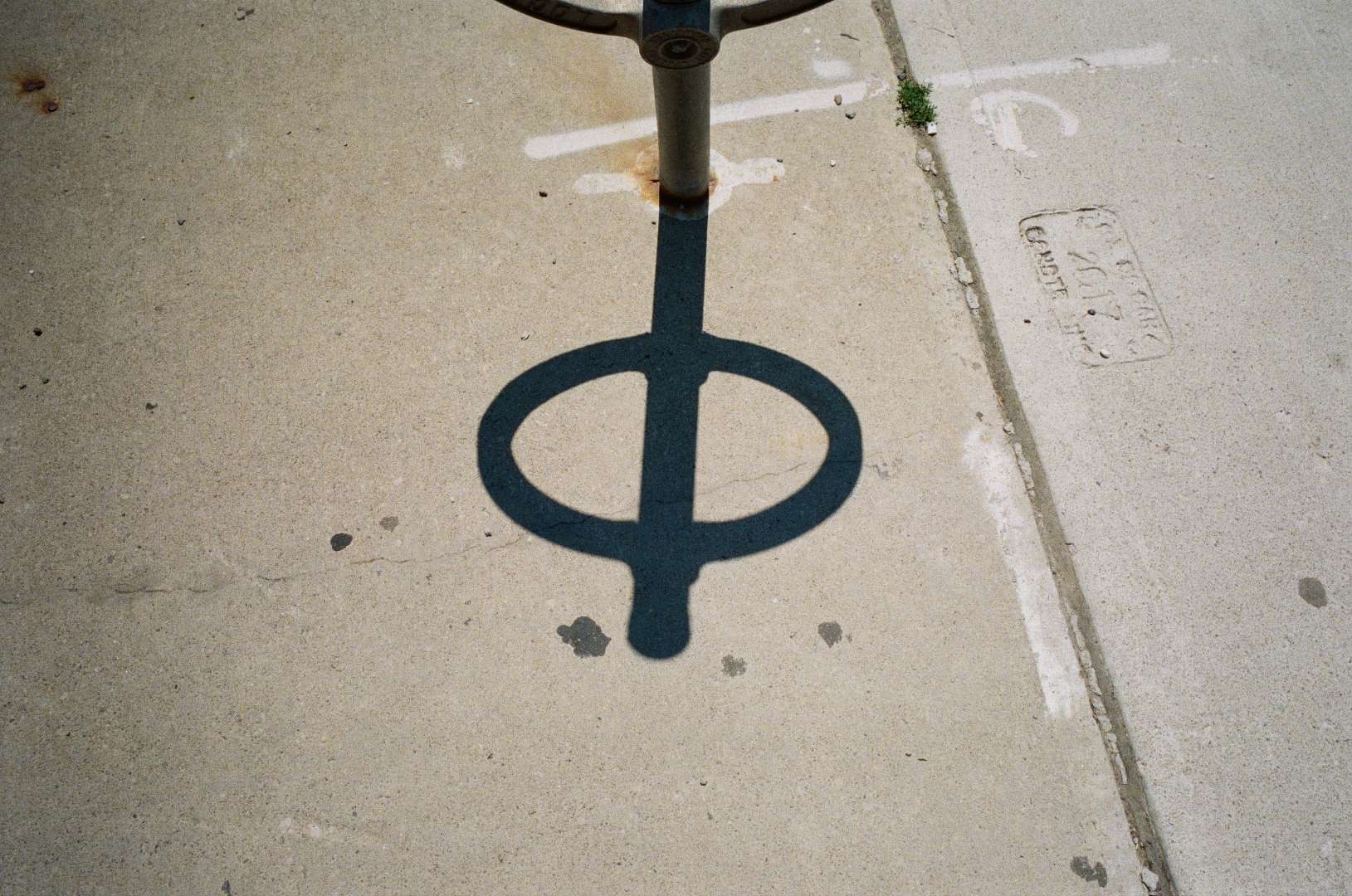A colour photograph of the shadow of a bike ring and post, the shadow marking time against stains on the sidewalk.