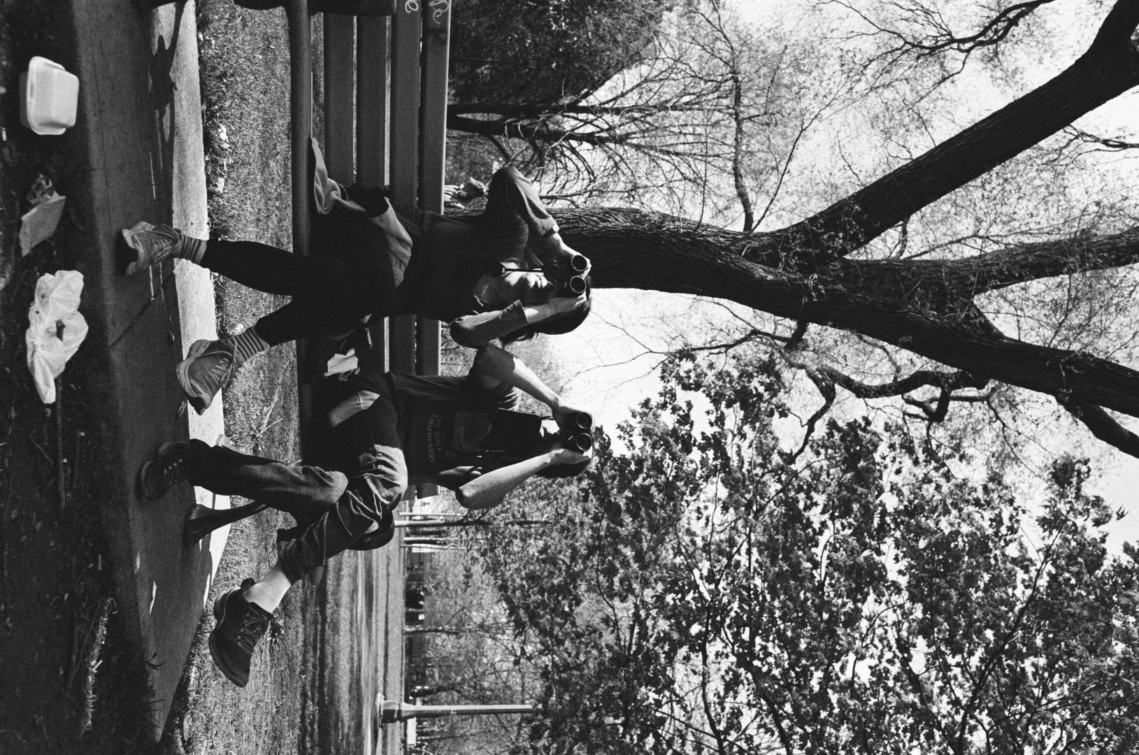 A black and white photograph of a man and a woman with binoculars held to their eyes, both have their legs crossed in the same way.